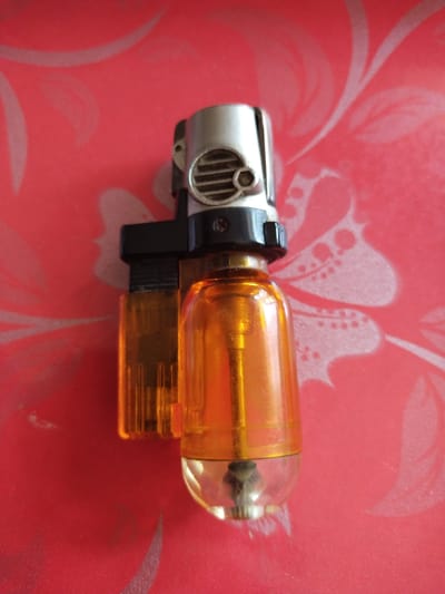 A lighter is positioned on a red patterned surface. The lighter has a silver top with ventilation slits and a black plastic component. The main body of the lighter is a translucent orange cylinder, revealing a metal wick inside. The surface it rests on is a vibrant red with a subtle, lighter red floral or leaf-like pattern. The image was taken from a top-down perspective, with the lighter positioned slightly off-center.