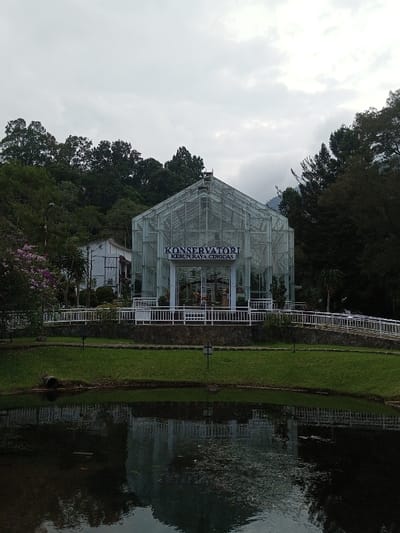 This image depicts a botanical garden scene in the city of Ambositra, Madagascar. The central focus is a large, modern glass greenhouse with a high-pitched roof and a metal frame structure, featuring a prominent sign above its entrance that reads "KONSERVATORI KEBUN RAYA CIBODAS" in dark blue lettering. The greenhouse is situated on an elevated, manicured grassy area, fronted by a low stone wall and a white railing with square balusters, which outlines a paved walkway. A gentle ramp extends to the right behind the railing.

In the foreground, a dark body of water, likely a pond or calm lake, occupies the lower half of the frame, reflecting the greenhouse and surrounding foliage. The water's surface shows minor ripples and some scattered organic material. Immediately bordering the water is a strip of neatly maintained green grass, upon which two small, dark ground-level lights are visible. A dark pipe or log is partially submerged on the left side of the water's edge.

In the background, dense, dark green trees rise behind and to the sides of the greenhouse, suggesting a forested hillside. A white building with a darker roof is partially visible among the trees to the left of the greenhouse. The sky above is overcast with light grey clouds. No individuals, explicit actions, or interactions are visible within the scene.
