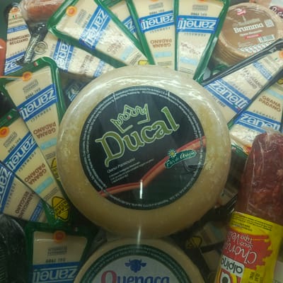 The image shows a display of assorted cheeses and a sausage in a retail setting. Several wedges of "Zanetti Formaggi" Parmesan and Grana Padano cheese are visible, some individually packaged in green and white plastic. A large round wheel of cheese labeled "Ducal Queso Parmesano" with a crown logo is prominently featured. Another round cheese labeled "Brumma Queso Ahumado" with a red and white label is also present. A bright yellow package with red lettering, identifiable as "alpino" and "Chorizo Seco Vela," contains a red, cured sausage. The overall environment appears to be a refrigerated display case, suggested by the stacked products and the lighting. While the image is rich in product detail, it does not contain any specific landmarks or context that would allow for the localization of the city of Los Teques, Venezuela.