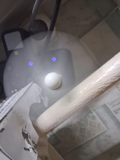 The image depicts a close-up, overhead view of a white, spherical object resting on a grey, circular electronic device with two illuminated blue power buttons. The device is covered in a layer of mist or vapor, suggesting it might be a humidifier or a similar appliance. A portion of a beige, textured cylindrical object, possibly a pipe or a support beam, is visible in the foreground, extending diagonally across the frame. The background shows what appears to be a tiled surface and a dark, possibly metallic, element. The overall scene is dimly lit, with the blue lights of the device and the white sphere being the most prominent sources of illumination. The image appears to be taken indoors, possibly in a residential or utility area. The location in Zhodzina, Belarus is not discernable from the visual information presented in the image.