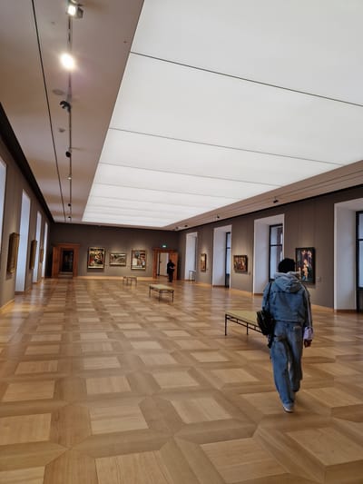 The image depicts an interior view of a long, rectangular art gallery hall within the Staatsgalerie at Schloss Johannisburg in Aschaffenburg, Germany. The hall features a polished light-brown wooden parquet floor arranged in a geometric, cube-like pattern. The walls are painted a neutral grey-brown and display multiple framed Old Master paintings.
In the foreground, a person is observed from behind, walking towards the distant end of the hall. This individual wears a light-blue denim jacket over a grey hooded garment, blue jeans, and light-colored shoes. They carry a black backpack on their left shoulder and a small light-purple bag in their right hand. Further down the hall on the right side, a second person, dressed in dark clothing, stands near a wooden doorway.
The ceiling features a combination of lighting elements: a track lighting system with multiple spotlights runs along the left side, and a large central rectangular panel provides diffused white light. Several framed paintings of various sizes are visible on both the left and right walls. The right wall also shows two elongated, backless benches with light-colored cushioned tops and dark metal frames. Architectural features include recessed vertical sections, possibly windows or niches with dark frames, along both side walls, and two ornate wooden doorways in the background, leading to other rooms.