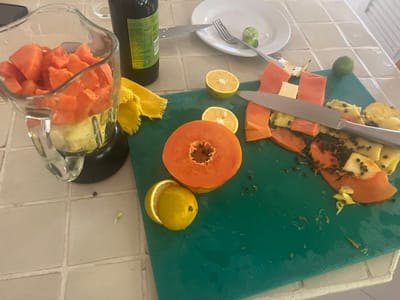A high-angle shot reveals smoothie preparation on a tiled surface. A blender with chopped papaya sits beside a green cutting board, which holds papaya halves, slices, pineapple pieces, and citrus fruits, including lemons and limes. A large knife rests on the cutting board. Other elements include a bottle, a plate, and a fork.