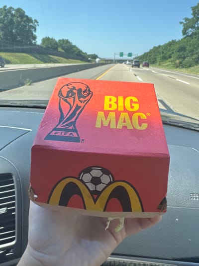 An adult's hand holds a red McDonald's Big Mac box with a FIFA World Cup trophy graphic and the text "BIG MAC" on one side. The box has a soccer ball graphic and McDonald's golden arches on the bottom. The scene appears to be inside a vehicle, with a dashboard visible, and the box is being held up against the backdrop of a highway on a sunny day. Trees and road signs are visible in the background. The location is not identifiable as Madison, United States of America.