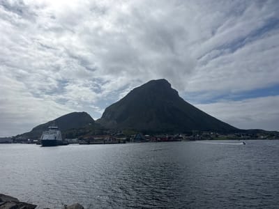 This image depicts the scenic coastal town of Lovund, Norway, under a cloudy sky. In the foreground, the dark, rippling water of the harbor fills the lower half of the frame. Rocky formations are visible along the immediate left edge of the foreground.
In the mid-ground, a large ferry boat is docked on the left side of the harbor. Behind the ferry, the town of Lovund is nestled along the shoreline, characterized by a cluster of red and white buildings with traditional Norwegian architecture. A prominent, dark, and imposing mountain dominates the background, rising steeply behind the town. Its slopes are partially covered in greenery.
Further to the right in the mid-ground, several smaller boats are visible on the water. One of these, a jet ski, is actively moving, leaving a white wake behind it. The sky above is a mix of bright white and gray clouds, with patches of blue sky visible between them. The overall impression is one of a calm, yet dramatic, coastal landscape.