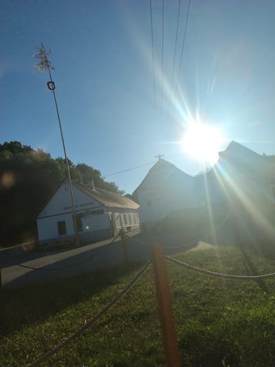 The image depicts an outdoor scene in Bošovice, Czechia, on a sunny day with a bright sun flare. In the foreground, there is a grassy area with a rope fence held up by wooden posts. Behind the fence, a paved area is visible, leading to a white building with a brown roof. Text on the building reads "Bosovská hospůdka". Further in the background, another building with a similar roof is visible, and overhead, power lines stretch across the sky. A tall wooden pole with some decorations at the top stands to the left of the white building. The sky is clear blue.