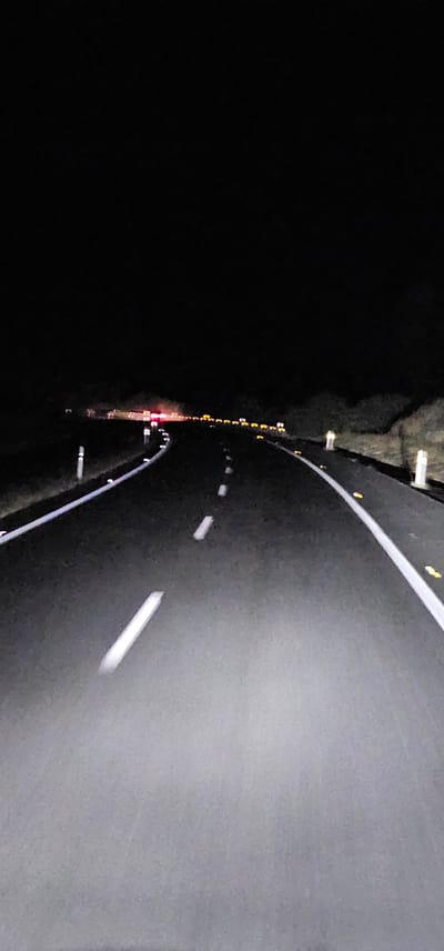 The image depicts a nighttime view of a highway in Santiago del Campo, Spain. The perspective is from the driver's seat, looking forward along the asphalt road. White dashed lines mark the lanes, and the road edges are delineated by white lines and reflective markers. In the distance, the headlights and taillights of other vehicles are visible, creating streaks of light against the dark landscape. The sky is completely black, indicating it is nighttime. The surrounding environment appears to be rural, with dark, unlit terrain visible on either side of the road. There are no visible people or specific landmarks that would identify this location within Santiago del Campo beyond it being a highway.