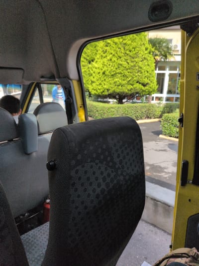 The image shows the interior of a yellow van, with a focus on the back seats and a view out the open side door. The seats are upholstered in dark fabric with a patterned texture. A person's head is visible in the left side of the frame, with dark hair. In the background, through the windows, there is a lush green tree and a building with white window frames. The ground outside appears to be paved, with some water on the surface. There is also a hedge and some potted plants visible. The overall scene suggests a daytime setting, likely outdoors in a temperate climate. There are no elements in the image that can identify its location in the city of Şəki, Azerbaijan.
