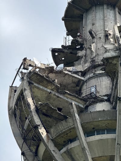 The image shows a dilapidated, concrete structure against a pale sky. The structure appears to be an unfinished or partially destroyed building in Sremska Kamenica, Serbia. The concrete is heavily damaged, with large chunks missing, exposing rebar and interior spaces. The architecture features a mix of curved and angular elements. Satellite dishes are visible, attached to the crumbling concrete. The overall impression is one of decay and abandonment. There are no people visible.