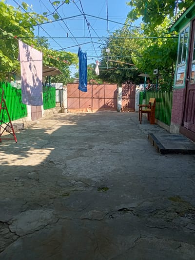 This is an outdoor image taken during the daytime in Salhany, Ukraine. The scene depicts a courtyard with a concrete ground that shows signs of wear and tear, with visible cracks and patches. A green corrugated metal fence runs along the left and back sides of the courtyard, with a large, reddish-brown metal gate in the background. A wooden bench is positioned near another green fence on the right side of the courtyard, adjacent to a brick building with a brown door and window. Overhead, a structure with wires and a grape vine provides shade. Laundry, including a pink towel and a blue towel, is hanging on clotheslines, attached with clothespins. A red ladder stands to the left of the frame. The overall impression is of a residential yard or an area behind a building. The sky is clear and blue.