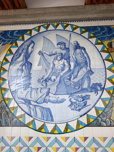 This is a tiled mural in Terras de Bouro, Portugal, depicting a scene of men fishing. The central circular motif showcases three men in a boat, hauling in a net filled with fish. One man, with a beard and wearing a cape, stands in the background. Another man, muscular and bare-chested, is actively pulling the net. A third man, wearing a cap, is also involved in the fishing activity. To the left, a figure with long, flowing hair, possibly a representation of Jesus, extends a hand, seemingly directing the fishermen. The water is depicted with waves, and various fish are scattered around the boat and in the net. The mural is bordered by a decorative pattern of triangles in blue, yellow, green, and white. The background outside the circle includes stylized clouds and hints of landscape, with a building or structure visible on the right. The bottom portion of the mural features a repeating geometric pattern of colorful tiles. The style is reminiscent of traditional Portuguese azulejos.