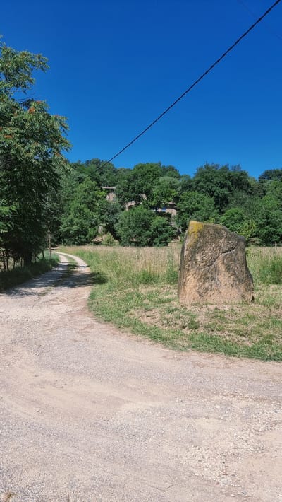 The image captures a scenic view in Montagut i Oix, Spain, featuring a dirt road winding through a grassy field under a clear blue sky. A large, weathered rock with inscriptions stands prominently in the foreground to the right. Dense green trees form a backdrop, with a glimpse of a stone building nestled within them. A power line stretches diagonally across the upper portion of the frame, adding a touch of modern infrastructure to the natural landscape. The overall impression is one of rural tranquility and historical resonance.