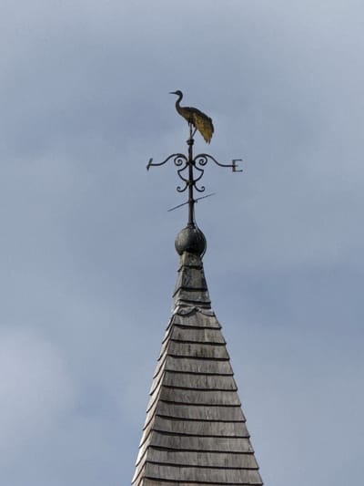 An upward, oblique shot shows a weather vane, likely situated atop a building with a steep, tiled roof. The vane features a bird-shaped figure, along with stylized directional indicators. The background is a muted, gray sky. The image suggests a location in Dacorum, United Kingdom, although specific details for pinpointing the exact location aren't included.