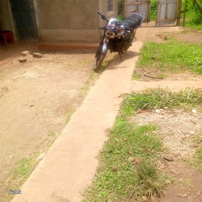The scene, located in Bonyamaro ward, Kenya, depicts an outdoor environment under daylight conditions. A black motorcycle is centrally positioned, parked on a concrete walkway that bisects the frame diagonally from lower left to upper right. The motorcycle, resting on its kickstand, displays a patterned black seat and features visible headlights with decorative elements. To the left of the concrete path, a dirt and gravel ground surface leads to a light-colored, single-story building with a visible window. A red plastic chair and several loose red bricks are positioned near the building's base. To the right of the walkway, patches of green grass and scattered vegetation are present. In the background, an open gate constructed from corrugated metal is visible, with dense green foliage beyond it. No persons are present, and no active interactions or movements are observed.