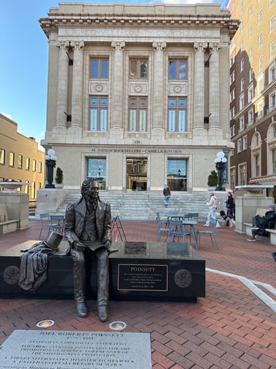 A bronze sculpture of Joel Roberts Poinsett sits on a black monument in front of Greenville County Courthouse at 130 M. Judson Booksellers • Camilla Kitchen. The building behind the statue is three stories tall with columns and detailed architectural features. The monument includes information about Poinsett's life as a statesman, diplomat, and naturalist, noting his role in establishing the Smithsonian Institution. Chairs sit behind the statue and monument, with people on the steps to a building.