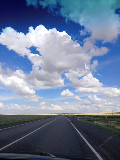 The image shows a view from inside a vehicle looking forward along a paved road. The road has white lane markings and extends into the distance, flanked by grassy fields and some vegetation. In the background, the landscape appears flat with a line of trees or poles in the distance. The sky is a deep blue with large, fluffy white clouds, and some lighter blue and turquoise colored clouds near the top of the frame. The overall scene depicts a rural or steppe landscape under a dramatic, cloudy sky. There are no people or identifiable structures that would indicate a specific city or location.