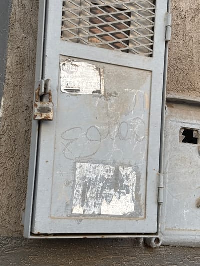 A close-up, eye-level, outdoor shot shows a gray metal utility box against a textured concrete wall. The utility box is unlocked and has a small, rusty padlock attached. On the front of the box, there is a partially peeled sticker with Arabic writing and a phone number. There is also some faint graffiti on the box that appears to be numbers or symbols. To the right of the main box, there is another compartment with a broken window. The overall condition of the utility box and its surroundings suggests it is located in an urban environment and may be in disrepair. The image was taken on a cloudy day, with the light appearing flat.