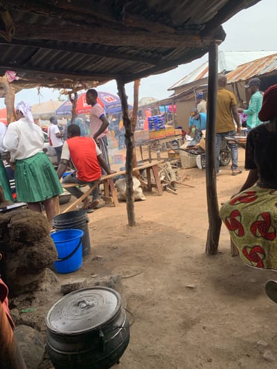 The image depicts an outdoor market scene in Dull A, Nigeria. Several people are present, some are standing, while others are seated. In the foreground, a large metal pot with a lid sits on the ground, possibly used for cooking. To the left, a blue plastic bucket and a black plastic container are visible. The environment appears to be dusty and rural, with makeshift structures providing shade from the sun. In the background, there are stalls with various goods displayed, including packaged food items. The overall atmosphere suggests a busy marketplace environment.