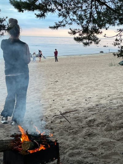 This image depicts a beach scene in Lebyazhye, Russia, with a bonfire in the foreground and a body of water in the background. A young woman is standing facing away from the camera, her back towards the viewer, with her hair in a bun. She is wearing a dark jacket and dark pants, with white sneakers. Smoke from the bonfire partially obscures her lower body. In the mid-ground, several other individuals are visible on the sandy beach. Two are walking towards the water, one in a dark swimsuit and the other in lighter clothing. Further back, a man in a burgundy jacket is standing near the water's edge. The water appears calm, with a pale horizon line suggesting it might be early morning or late evening. The beach is sandy and appears to have some scattered driftwood and natural debris. Pine branches overhang the scene from the top, framing the view. The overall atmosphere is relaxed and casual, indicative of a beach outing.