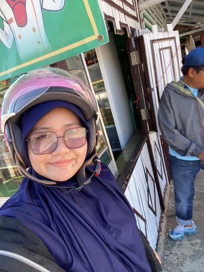 A woman wearing a dark blue hijab and helmet, with clear-lensed glasses, smiles at the camera. She is in the foreground, positioned on the left side of the frame. Her dark blue garment covers her head and shoulders. Behind her, a man wearing a blue baseball cap, gray jacket, and blue jeans is visible. He is standing near a building with white paneled walls and brown trim. The building appears to be a shop or market, with a green banner displaying a cartoon image visible above the woman. The ground is covered in a light-colored substance, possibly gravel or debris. The overall scene suggests an outdoor setting in a public area.