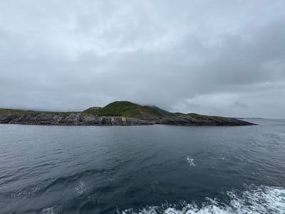 An overcast, grey sky looms above a body of dark, choppy water. In the middle ground, a long, narrow island stretches horizontally across the frame. The island's foreground is characterized by rugged, grey rock formations, interspersed with patches of green vegetation. The island's interior rises into gently sloping hills covered in lush green foliage, with some sections obscured by mist. The water in the foreground exhibits white foam and ripples, suggesting movement. The overall scene depicts a coastal landscape under a cloudy sky in Demmahågen, Norway.