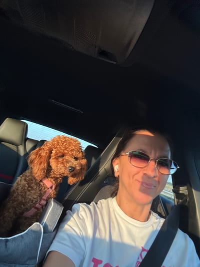 The image shows a woman and a toy poodle in a car. The woman is in the driver's seat, wearing a white t-shirt, sunglasses, and earbuds. She is smiling and looking at the camera. The poodle is in a pet car seat in the passenger seat, wearing a pink harness. The car's interior is visible, including the headrests and seatbelts. The background shows a road with trees and a body of water, suggesting the car is driving through a rural area. The scene appears to be a selfie taken by the woman. There are no visible indications that this image was taken in Scheemda, Netherlands.