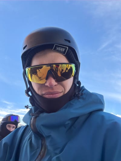 The scene is an outdoor winter environment in Lunnaya Polyana, Russia, under a clear blue sky with scattered white clouds. Two individuals are present, dressed in cold-weather gear. The primary subject is centered in the foreground, facing the camera directly, suggesting a selfie. This individual wears a black helmet, beneath which a black beanie with a white "VANS OFF THE WALL" patch is visible. Mirrored yellow-gold sports sunglasses cover the eyes, reflecting a snowy landscape, what appear to be other individuals, and vertical structures consistent with a ski resort. A black neck gaiter covers the lower face, and a teal-blue hooded winter jacket with a dark-colored waterproof zipper is worn. The subject's lips are slightly pursed. Partially visible in the lower-left background is a second individual, wearing a black balaclava that covers the head and most of the face, blue-purple reflective ski goggles, and a dark jacket. The second individual's visible eyes are directed towards the general camera position.