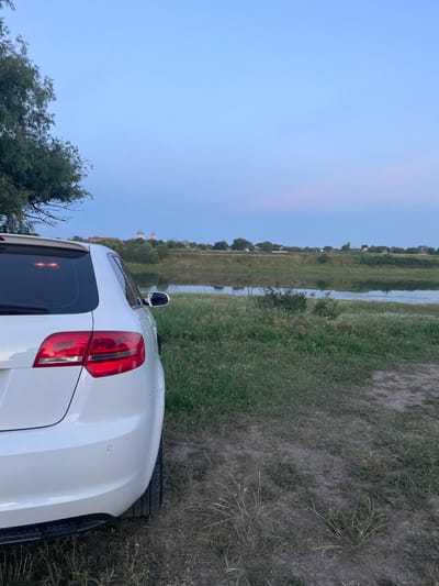A white car is parked on a dirt path next to a grassy area. The car is facing away from the viewer, with its rear end visible. In the background, there is a body of water, likely a river, with a line of trees and buildings on the opposite bank. The sky is a pale blue with hints of pink near the horizon. The scene appears to be in a rural setting, possibly on the outskirts of Coșnița, Moldova. The lighting suggests it is either early morning or late evening. There are no people visible in the image.