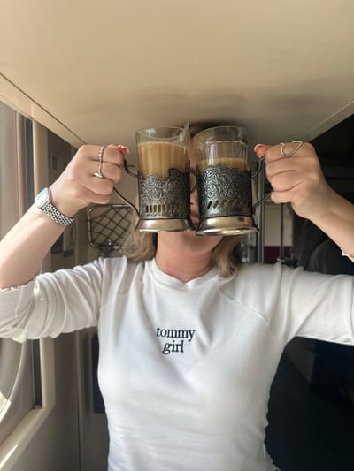 This close-up shot features a person, likely female, holding two clear glass cups in ornate silver-toned metal holders (podstakanniki). Each cup contains a light brown, milky beverage, possibly coffee or tea. The person is wearing a white long-sleeved t-shirt with the text "tommy girl" visible on the chest. Rings adorn her fingers, including a distinctive heart-shaped one on the left hand and a beaded one with a small pointed design on the right. A silver-toned wristwatch is visible on her right wrist.
The setting appears to be an interior space, characterized by beige walls and what looks like a train window or grille in the background, suggesting a journey. The cups themselves are traditional Russian podstakanniki, commonly used on trains. The overall lighting is bright, indicating it is daytime. The provided location context, Куркачи, Russia, further reinforces the likelihood of this being a scene from a Russian train journey. The person's face is mostly obscured by the cups, so specific emotions are not discernible.