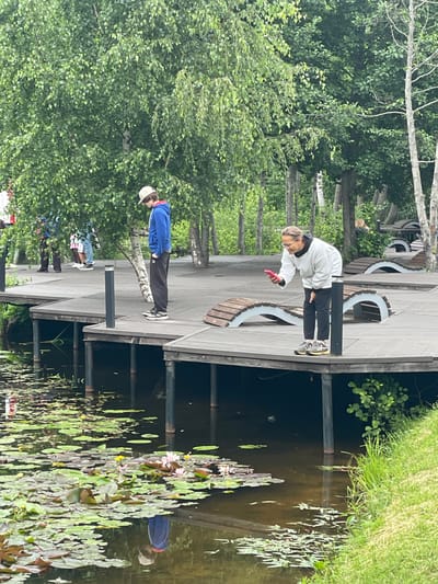 The media file shows an outdoor scene on a wooden boardwalk next to a body of water filled with water lilies. There are three people visible on the boardwalk. The person in the foreground is wearing a white hoodie and black pants and is looking down at a pink object, possibly a phone, in their hand. To their left and slightly behind them, another person is wearing a blue jacket and a light-colored cap, with black pants. Further down the boardwalk, a third person is visible from the waist down, wearing dark pants and dark shoes, with some colorful clothing items peeking out from behind them.

The setting appears to be a park or recreational area, with trees and lush green foliage surrounding the boardwalk. The water is dark and reflects the wooden structure and surrounding vegetation. The presence of water lilies suggests a natural or semi-natural aquatic environment. The lighting indicates it is daytime, and the weather appears to be overcast or partly cloudy, given the diffused light and lack of strong shadows. There is no visible text in the image. The overall mood is casual and relaxed, with individuals enjoying the outdoor setting. The location context provided is Sosnoviy Bor, Russia.