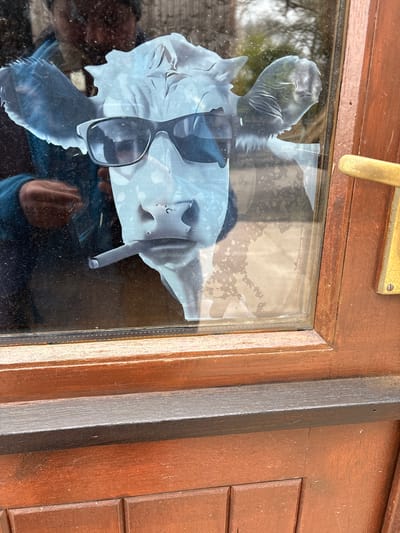 A close-up of a brown wooden door with a rectangular glass pane. A distorted image is stuck on the inside of the pane. The image features an incomplete impression of a human face and the shape of a pair of sunglasses. The environment is not discernible and the location in Pulverbatch, United Kingdom is not determinable from the image.