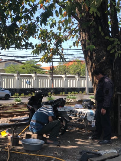 In Buduran, Indonesia, a man in a dark jacket and pants stands near a tree, looking down. Another man, wearing a blue and white striped shirt and dark pants, is crouched near a motorcycle, working on it. A motorcycle is parked on the side of a road with train tracks. In the background, a white fence with pillars lines the street, and buildings with orange roofs are visible. The scene is bathed in sunlight, with shadows cast by the tree and foliage overhead.