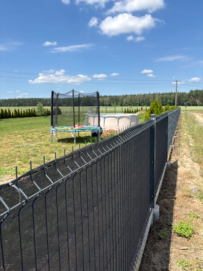 The image depicts a rural backyard scene in Opatów, Poland, on a clear day with a blue sky dotted with white clouds. A dark, mesh fence runs diagonally across the foreground, separating the viewer from the backyard. Beyond the fence, a green lawn is visible, featuring a trampoline with a safety net and a blue frame, and a large, round above-ground swimming pool. A line of manicured evergreen trees borders the lawn in the mid-ground, with a dense forest in the background. A utility pole with power lines stretches across the sky. The fence appears to be a newly installed privacy fence, indicated by the concrete base and the plastic screening within the wire mesh. The ground alongside the fence is a mixture of dry grass and dirt.