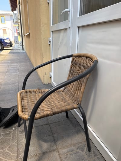 The image shows an outdoor scene, likely a patio or balcony, with a wicker chair as the central object. The chair has a dark metal frame and a woven seat and backrest. The chair is positioned in front of a white wall and a window, with grey tiled flooring beneath it. In the background, there is a glimpse of an alleyway with a building, a blue car, and some overhead lighting fixtures, suggesting an urban or suburban environment. The lighting indicates it is daytime, with sunlight casting shadows on the floor. The overall scene is mundane, depicting an everyday outdoor setting. There is no visible text in the image.