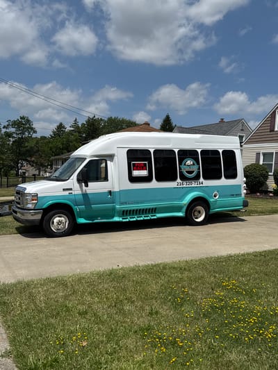 The image shows a white and teal passenger van parked on a concrete driveway next to a well-maintained lawn with scattered yellow flowers. The van has several windows along its side, some tinted. A "FOR SALE" sign is visible in one of the windows. Text on the van indicates "WRIGHT CHOICE TRANSPORTATION" with a phone number "216-320-7184" and a website "www.wrightchoicetransportation.com". The van also has the text "NON-EMERGENCY" printed on its side. In the background, a residential house with a brown and white exterior is visible, along with some trees and a power line. The sky is blue with scattered white clouds, suggesting it is a sunny day. The overall scene depicts a vehicle for sale in a suburban setting. The image is oriented vertically.
