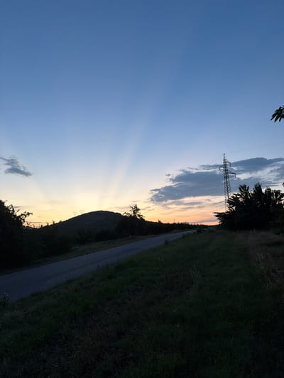 The image depicts a landscape scene in Shatrovo, Bulgaria, likely during the late afternoon or early evening, as indicated by the soft, warm glow of the setting sun on the horizon and the crepuscular rays of light emanating from behind a silhouetted hill. The sky transitions from a clear, deep blue on the left to lighter shades of yellow and orange near the horizon.
A prominent feature of the scene is a tall, metal electricity pylon with cables stretching across the frame, suggesting the presence of power infrastructure. To the right of the pylon, a dense line of dark green trees creates a stark contrast against the brighter sky. Beyond the trees, a road or path is visible, running parallel to the tree line and disappearing into the distance. The ground to the right of the road is covered in dark green grass, suggesting a rural or semi-rural environment.
A distinct silhouetted hill dominates the middle-ground, with its dark contours sharply defined against the illuminated sky. Subtle shadows and the texture of the terrain on the hill are visible, though details are obscured by the low light conditions. A few small, dark clouds are scattered across the sky. The overall mood is serene and peaceful, with the natural beauty of the landscape enhanced by the dramatic lighting of dusk. There is no visible text in the image.