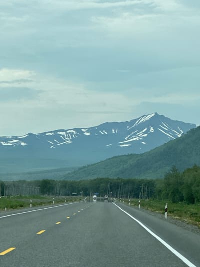 The image shows a paved road with a white dividing line and yellow dashed lines in the foreground, leading towards a vast mountain range in the background. The mountains are covered with patches of snow, indicating a high altitude or a cold climate. The sky is overcast with light gray and blue clouds. Along the sides of the road, there are utility poles and vegetation, suggesting a rural or semi-rural setting. The scene appears to be a landscape photograph taken from a moving vehicle, as evidenced by the perspective and the motion blur on the road markings. There are no people or animals visible in the image. The location is specified as Кеткино, Russia.