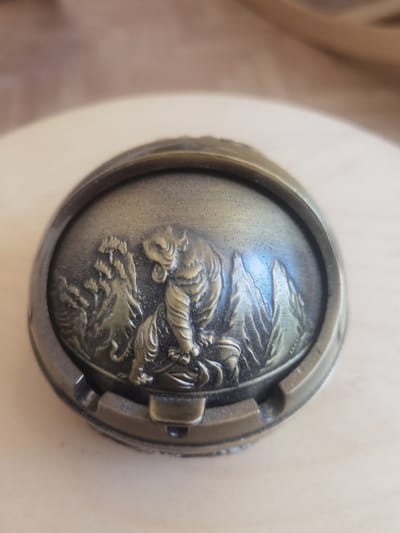 The image shows a close-up, eye-level shot of a decorative, oval-shaped object with a metallic finish, possibly an ashtray or a lid. The object is placed on a light-colored, smooth surface, which appears to be a wooden table. In the background, a blurred, wooden surface is visible. The central subject is an embossed design depicting a tiger in a natural setting. The tiger is positioned in the center of the oval, appearing to be walking or prowling. It is facing towards the viewer's left, with its head turned slightly. Behind the tiger, there are representations of rocky terrain and some vegetation, possibly trees or bushes, adding depth to the scene. The metallic material of the object has a bronze or antique gold color, with some darker areas suggesting patina or shadows. The design is detailed, showcasing the musculature of the tiger and the textures of the landscape. The overall scene is static, focusing on the craftsmanship of the object and its embossed imagery. There is no visible text in the image. The lighting appears to be from above and to the left, casting shadows and highlighting the raised areas of the embossed design. The focus is sharp on the object, with the background intentionally blurred.