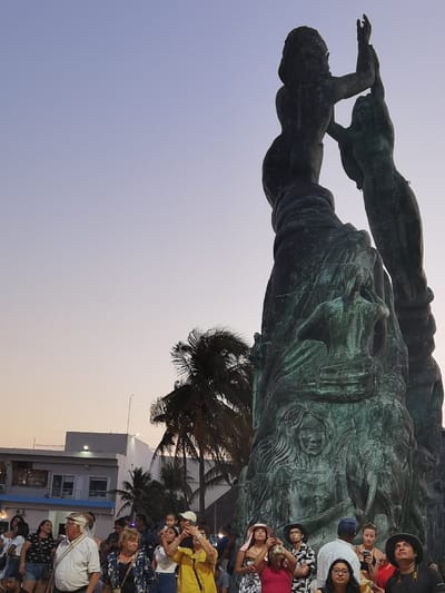 The scene shows a large bronze statue of two women, located at the Parque Benito Juárez in Cozumel, Mexico (latitude: 20.509, longitude: -86.949). The statue is situated in a plaza, surrounded by palm trees and a few buildings behind. There are several people around the statue, looking up at it and taking pictures. Some are holding cameras and some are holding cell phones. There is a group of people near the front of the statue, with several children among them. Some people are wearing hats. The sky is a pale blue, suggesting late afternoon or early evening. The people are likely tourists visiting the city.