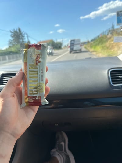 A person's hand holds a protein bar from Prozis, with the bar's wrapper prominently displayed. The wrapper is primarily white and gold, featuring the brand name "Prozis" and the product name "NO 1 BILLION." There's an image of a dessert-like bar on the wrapper and text indicating "WHITE CHOCOLATE BAR" and "PEANUT BUTTER." A circular emblem reads "ONE IN A BILLION" and "20g PROTEIN." The bar is 68g.
The scene appears to be inside a car. The dashboard and a vent are visible. Outside the car, a bright, sunny day is suggested by the clear blue sky and strong sunlight casting reflections on the windshield. Green foliage and a road with other vehicles are visible through the windshield, indicating the car is in motion. The background suggests a rural or suburban setting.
The person's hand holding the bar is relaxed, and the overall mood seems casual and perhaps on-the-go, given the context of being in a car. The time of day is likely midday due to the bright sunlight. The location context of Alès, France, is provided but not directly evident from the visual cues.