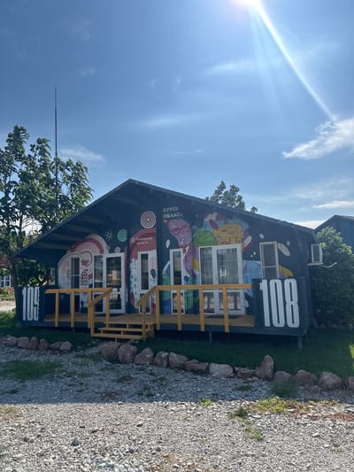 A dark blue, single-story building with a pitched roof and a yellow-railed porch is decorated with a vibrant mural and the number "108" in large white font on both ends of the facade. The mural features stylized illustrations of people, architectural elements, and abstract shapes. The building is situated in an outdoor environment with a gravel driveway and scattered rocks in the foreground, and trees and a clear blue sky with a bright sun and wispy clouds in the background. The image appears to be taken on a sunny day. There is no indication of the city of Sudak, Ukraine in the image.