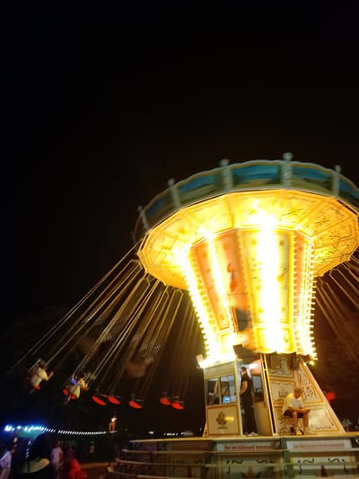 This image captures a scene at an amusement park at night, possibly at the Stadtfest in Cottbus - Chóśebuz, Germany. The central focus is a brightly illuminated swing ride, its yellow and blue canopy glowing against the dark sky. Suspended from the ride's mechanism are numerous chains, each holding a swing seat with people seated in them, appearing as colorful blurs due to the motion and low light. The ride's central structure is octagonal and decorated with intricate patterns and lights.
At the base of the ride, there is a small ticket booth or control station. Two individuals are visible near this booth. One person, wearing a dark t-shirt and pants, stands inside the booth, seemingly attending to operations. Another person, in a white t-shirt and shorts, is seated on a chair outside the booth, looking down at something in their hands.
In the background, blurred lights suggest other attractions or stalls within the park. Further down, a line of more lights, possibly decorative, stretches across the scene. The ground level of the ride's platform features seating areas with benches and tables, and some promotional signage is visible. The overall atmosphere is one of a lively, nocturnal fairground experience.