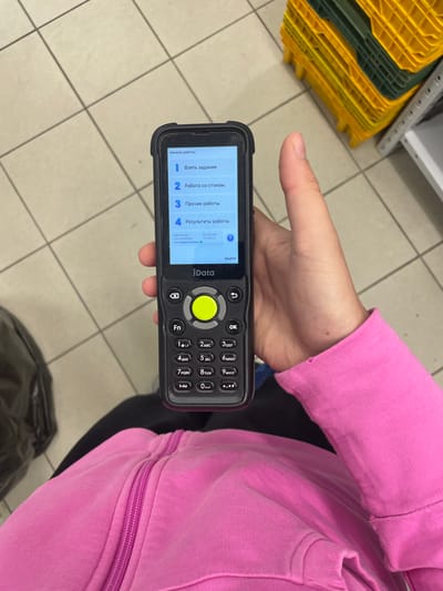 A hand wearing a pink hooded sweatshirt is holding a handheld scanner with a touchscreen display and a physical keypad. The scanner displays a menu in Russian with options for "Start Work," "Take Task," "Work with Stock," "Other Tasks," and "Task Results." The environment appears to be a retail or warehouse setting, indicated by the tiled floor and yellow and green plastic crates in the background. The scanner's screen shows user information and version numbers. The overall scene suggests a worker using a device for inventory management or task tracking.