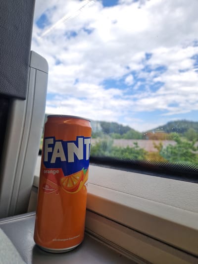 The image shows a can of Fanta Orange placed on a window ledge inside a moving vehicle, likely a train, as indicated by the reflection of overhead wires on the window. The can is predominantly orange and blue, with the "FANTA" logo clearly visible. A graphic of an orange slice is also depicted. Text on the can reads "orange" and "Mit Orangensaft" (with orange juice). The beverage is likely in Germany, specifically Radolfzell am Bodensee, based on the provided location context.

The scene outside the window depicts a bright, partly cloudy sky with fluffy white clouds against a blue background. Green foliage and hills are visible in the distance, suggesting a daytime setting, possibly late morning or early afternoon, given the lighting. The clouds and clear sky suggest fair weather. The interior of the vehicle appears to be a neutral beige color, with textured surfaces. The can is positioned at an angle, with the label facing the viewer. The overall mood is casual and relaxed, perhaps depicting a moment of travel or leisure.