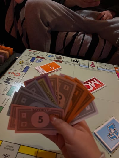The image captures an indoor scene within the Gerakas Municipal Unit, Greece, depicting a board game of Monopoly in progress. In the lower-center foreground, a human hand holds a fanned array of multi-colored Monopoly banknotes, including denominations such as a pink '5' note. The central and left portions of the frame are dominated by the Monopoly game board, which displays Greek text on its property and action spaces. Visible on the board are a stack of orange "Chance" cards, each marked with a white question mark, and a stack of blue "Community Chest" cards featuring a white treasure chest icon in the lower-right. A silver-colored thimble game token rests on one of the board's property spaces. The red "MONOPOLY" logo is visible on the game board. In the upper-central background, the lower torso and legs of a second individual are visible; this person is seated with their legs crossed, wearing light grey sweatpants with black stripes and a dark upper garment. A black container, partially obscured on the far left, holds additional Monopoly money. The environment is illuminated by warm, ambient lighting.