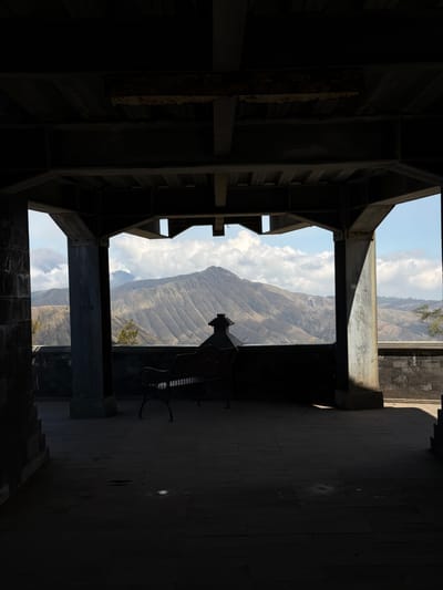 The image captures a view of volcanic mountains from a covered viewing area in Cemorolawang, Indonesia. In the foreground, a dark, open-sided structure with a tiled floor and a dark metal roof creates a silhouetted frame. Within this structure, a dark metal bench with a decorative backrest is visible, facing the panoramic mountain landscape. The mountains themselves are layered and rugged, with visible erosion patterns and some sparse vegetation. The sky is partly cloudy, with bright white cumulus clouds contrasting against a clear blue sky. The lighting suggests it is daytime, with strong shadows cast by the structure onto the floor. The overall scene conveys a sense of stillness and natural beauty viewed from a man-made vantage point.