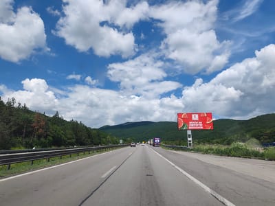 The image shows a view from a highway in the direction of travel, with a bright blue sky and fluffy white clouds overhead. On the right side of the highway, there is a large billboard advertising Kaufland with images of watermelons and the text "ВКУСЪТ НА ЛЯТОТО" (The taste of summer). The highway is bordered by a metal guardrail on the left and lush green vegetation and trees on the right, leading up to rolling hills and mountains in the background. Several vehicles, including cars and trucks, are visible on the highway ahead. The weather appears to be clear and sunny. The location is identified as Potop, Bulgaria.