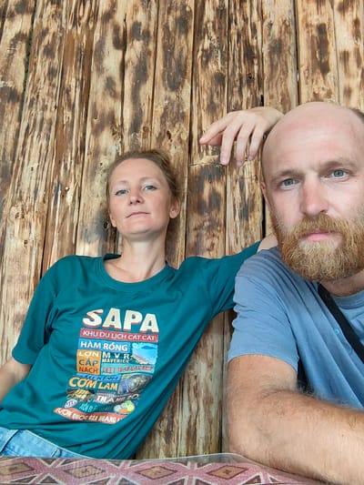 A man and woman are sitting close to each other in what appears to be a restaurant setting in Thành phố Hà Giang, Vietnam. They are posing for a photo, leaning against a wall constructed of rough, unfinished wooden planks. The woman is wearing a green t-shirt featuring a graphic design with the word "SAPA" prominently displayed at the top, followed by other text and images in Vietnamese relating to tourism in the region. The man is wearing a blue t-shirt. He has a shaved head and a red beard, and his arm is resting on the table in front of him. The table has a geometric patterned covering. The environment suggests they are in a rustic, possibly rural, location.