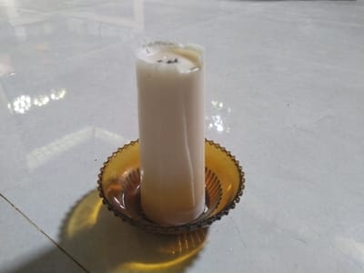 The image features a white, cylindrical candle resting in an amber-colored glass holder. The candle shows clear signs of previous use, with its top portion melted and uneven, revealing dark specks where the wick once burned. Some solidified, yellowish wax has accumulated in the bottom of the holder around the candle's base.

The holder is a decorative, shallow bowl made of amber glass, characterized by a distinct scalloped or beaded rim and a ribbed texture on its interior base. This holder casts a subtle yellow reflection and shadow on the surface it rests upon.

The candle and holder are placed on a light-colored, possibly tiled or concrete, floor or flat surface. This surface exhibits a slight sheen, reflecting blurred bright spots that suggest artificial overhead lighting or reflections from nearby windows. A faint, thin dark line, resembling a grout line or seam, crosses the surface in the lower left quadrant.

The scene is a static still life, implying an indoor setting. There are no people visible, and no specific activity is taking place beyond the evidence of the candle having been burned. The image is captured with an overhead, slightly oblique perspective. No discernible text is present in the photograph. The overall impression is one of a simple, everyday object in a functional setting, likely indoors in Teupinbayu, Indonesia.