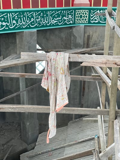 The image depicts the interior of a building under construction, identified as a mosque, located in Kumbang, Indonesia. The foreground and midground are dominated by a robust wooden scaffolding structure, composed of unpainted, rough-hewn timbers secured with visible nails. Draped centrally over a horizontal scaffold beam is a light-colored fabric, possibly a towel or cloth, patterned with small floral or cartoon designs in various colors, and featuring a pink trim along one edge.

In the upper background, above the scaffolding, a decorative green band spans the width of the image, featuring prominent white Arabic calligraphy and a geometric Islamic pattern. Above this green band, a red section with vertical brown stripes is visible. The background reveals unfinished concrete walls and columns. Windows composed of square glass blocks are visible through the scaffolding at multiple points. The lower background shows stacked gray rectangular tiles or slabs on the ground floor, alongside piles of construction debris, indicating ongoing work. No individuals are present in the scene.