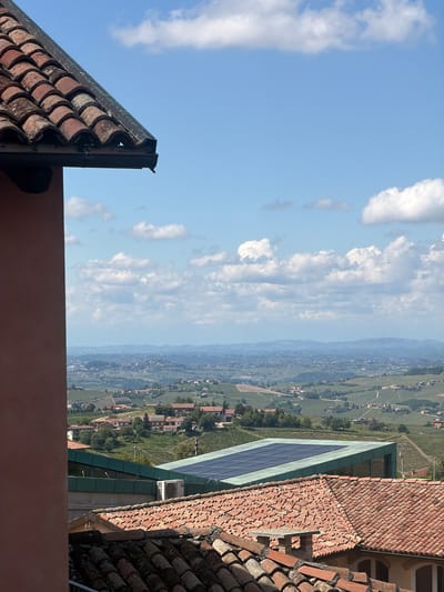 The image depicts a view of the Italian city of Castiglione Tinella, taken from an elevated perspective, likely a rooftop. The foreground includes sections of tiled roofs, typical of Italian architecture. A solar panel is also visible on a neighboring building. Beyond the roofs, the scene opens up to a wide landscape with a valley. Patches of green fields and vineyards are interspersed with houses and small structures, indicating a rural or semi-rural setting. The sky is blue with scattered clouds, contributing to a bright and sunny ambiance.